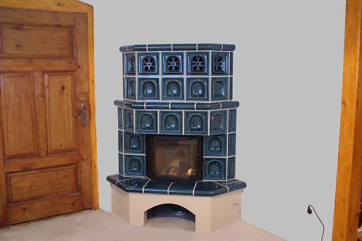 Kaflowy narożny piec z paleniskiem, obudowany ciemnozielonymi kaflami z dekoracyjnymi motywami, ustawiony w rogu pomieszczenia obok drewnianych drzwi i fragmentu drewnianej belki stropowej.